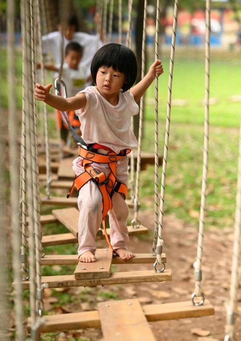 A young child carefully navigating a rope bridge in a harness, symbolizing courage, adventure, and personal growth encouraged by Rise, Roar, and Soar.