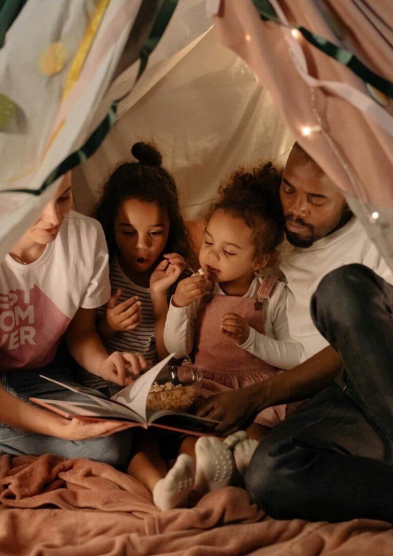A family of four sits together in a cozy tent, reading a book and sharing popcorn, symbolizing connection, mindfulness, and shared adventures.