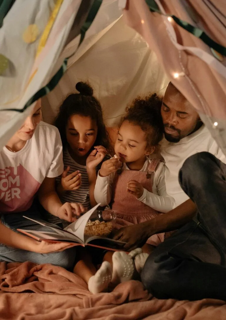 A family of four sits together in a cozy tent, reading a book and sharing popcorn, symbolizing connection, mindfulness, and shared adventures.