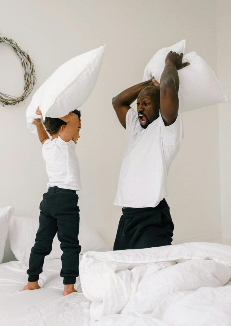 A joyful father and child playfully engaging in a pillow fight on a white bed, capturing a moment of connection, fun, and shared laughter.