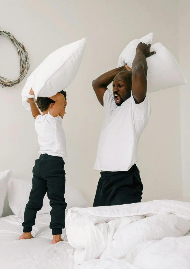 A joyful father and child playfully engaging in a pillow fight on a white bed, capturing a moment of connection, fun, and shared laughter.