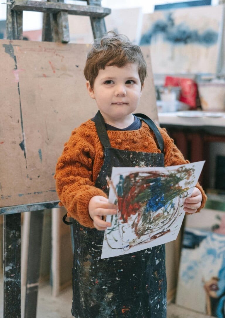 A young child proudly holding a colorful painting in a creative studio, symbolizing artistic expression and mindfulness activities found on Rise, Roar, and Soar.